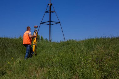 Professional Male Land Surveyor Measures Ground Control Point Using a GPS Rover. Green Field on a Background clipart