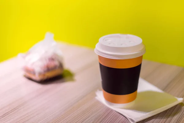 plastic cup of coffee is on the napkin. next to it is a wrapped hamburger. bright green yellow background.
