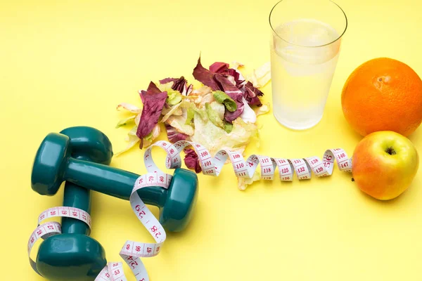 Dumbbells for sports. Fitness salad and tape measure on a yellow table. Oranges, apples and clear water with ice for the concept of a healthy lifestyle.