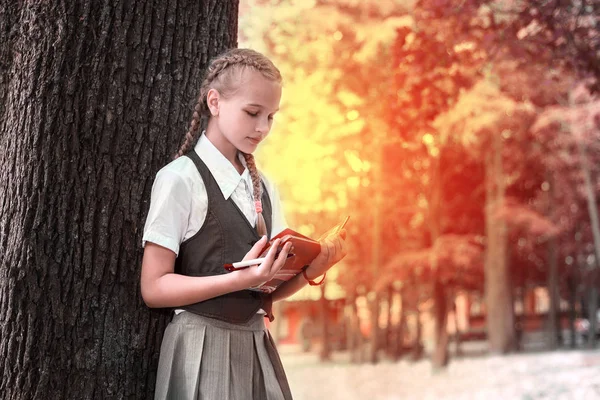 Colegiala adolescente leyendo un libro en un parque cerca de un árbol . — Foto de Stock