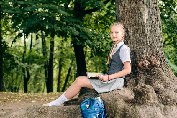 Colegiala adolescente leyendo un libro en un parque cerca de un árbol . — Foto de Stock