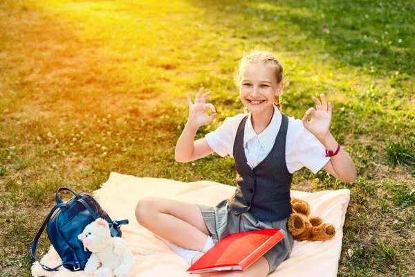 Colegiala alegre sonriendo y mostrando buena señal. el estudiante está sentado en un parque sobre una manta con juguetes de juguete blando que el oso sostiene — Foto de Stock