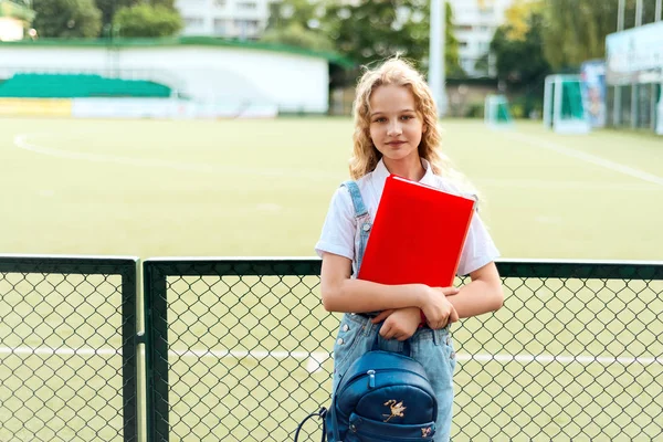 Μαθήτρια ξανθιά με μπλε μάτια κρατώντας ένα κόκκινο φάκελο και ένα σακίδιο. — Φωτογραφία Αρχείου