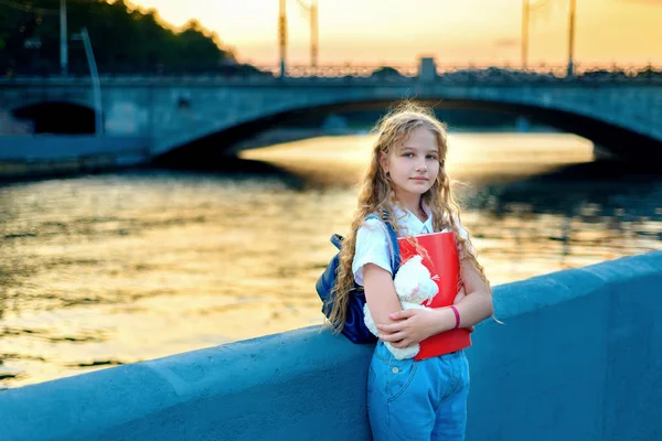 Colegiala rubia con ojos azules está de pie cerca del río en la ciudad al atardecer . — Foto de Stock