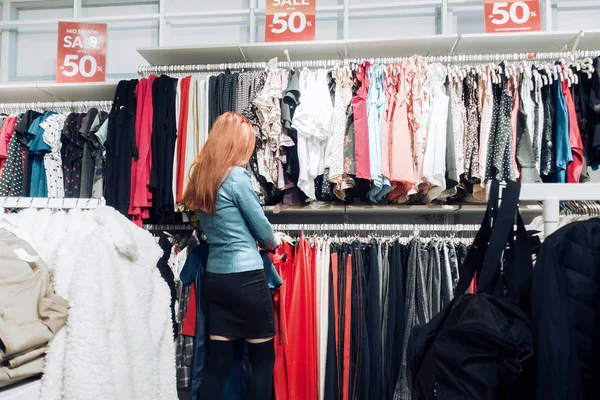 Chica joven de pelo rojo en una chaqueta de cuero azul elige la ropa en una tienda . —  Fotos de Stock