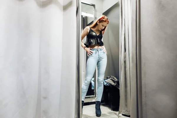 Chica joven pelirroja probándose unos vaqueros nuevos en el probador de la tienda. mujer se mira a sí misma en el reflejo del espejo. compra — Foto de Stock