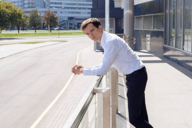 Tişörtlü ve pantolonlu genç adam binanın korkuluklarının yanında duruyor.