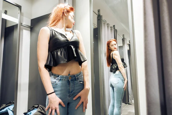 Червоне волосся молода дівчина намагається на нових джинсах у примірювальній кімнаті в магазині. жінка дивиться на себе в відображенні дзеркала. покупка — стокове фото