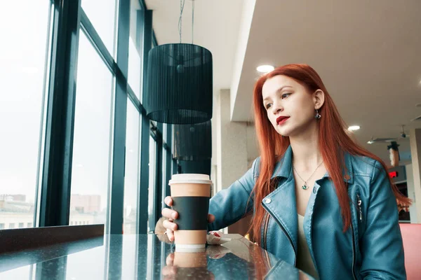beautiful lonely young girl in a cafe drinks coffee. pretty red-haired girl thoughtful
