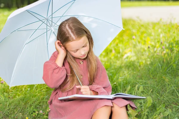 Ребенок читает книгу. Маленькая девочка под зонтиком сидит на зеленой траве в солнечный день — стоковое фото