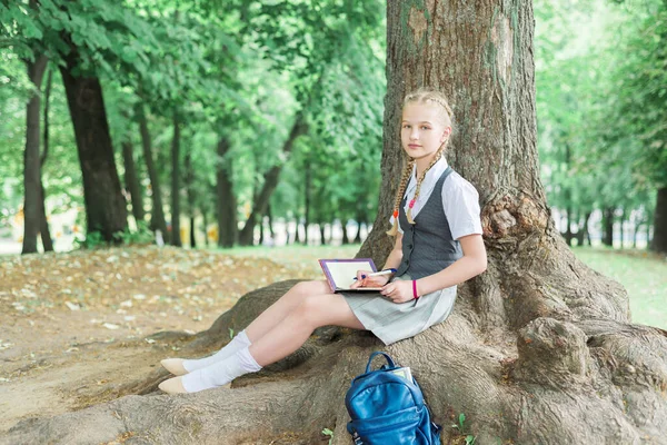 Colegiala enseña tareas en el parque cerca de un árbol grande — Foto de Stock