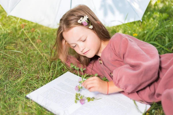 Милая маленькая девочка читает книгу. ребенок с цветами лежал на траве — стоковое фото