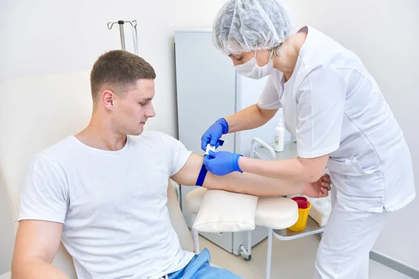 Tıp bürosundaki adam kan veriyor. Doktor koluna turnike yapıyor.