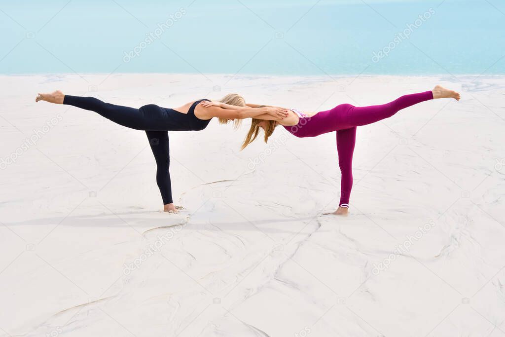 Two beautiful young women perform pose yoga virabhadrasana on the beach