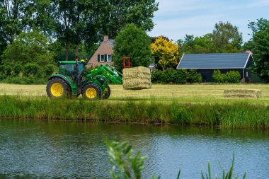 Dordrecht, Hollanda - Haziran 12020: Kırsal bölgedeki bir karavana balya saman yükleyen yeşil traktör. Biesbosch Ulusal Parkı Hollanda 'nın en büyük ulusal parklarından biridir.
