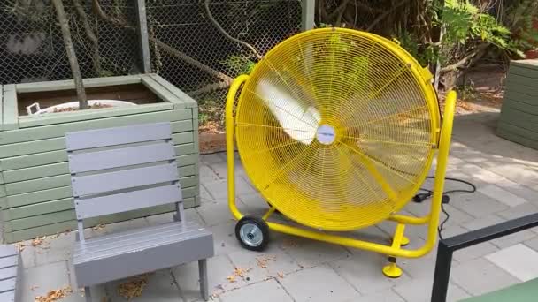 Large yellow industrial ventilator outside in a garden of a cafe restaurant. Keeping cool by the fan, air displacement, and adjustable airflow. Cooling down in the summer heat, blades turning fast blowing out cold air creating a breeze summer evening — Stock Footage Large Yellow Industrial Ventilator Garden Cafe Restaurant Keeping Cool Fan Royalty Free Stock Footage