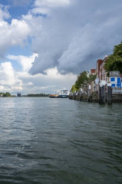 Dordrecht, Hollanda, 5 Eylül 2025: Fırtınalı gökyüzünün altında Oude Maas nehri boyunca eski tarihi Hollanda binaları yükseliyor, iç gemi ve bir yolcu gemisi yakınlarda demirliyor.