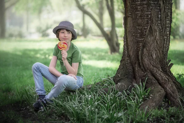 一个穿着 T恤和帽子坐在树根上的男孩的肖像 在他的手中一个甜美的糖果 — 图库照片