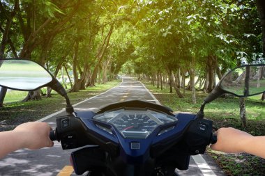 Hands of motorcyclist scooter on the road with trees. clipart