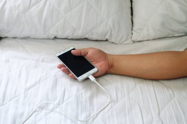 El kullanarak holding cep telefonları vardır. Beyaz yatakta şarj cihazı takılı