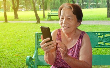 Yaşlı Asyalı kadın parkta bir gülümseme ile cep telefonu baskı yapıyor