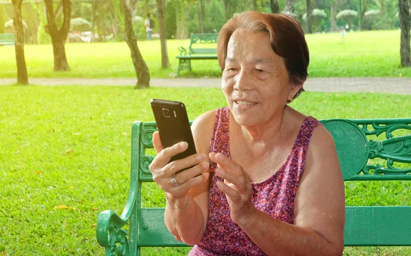 Yaşlı Asyalı kadın parkta bir gülümseme ile cep telefonu baskı yapıyor