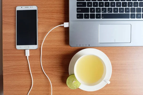 Plug in USB cord charger of the mobile phone with a laptop computer and freshly Lime juice in a white cup, on wooden floor, Top view