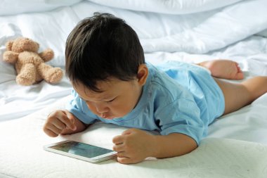 2 yaşında Asyalı bir çocuk, beyaz yatakta cep telefonu izliyor ve oynuyor..