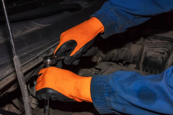 Gloved hands of mechanic holding wrench or spanner working on car engine. Mechanic wearing orange gloves and blue outfit covered with oil.