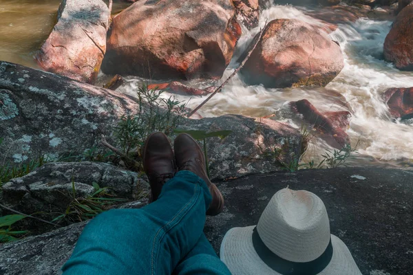 man feet sitting on stone at the riverside with brown shoes,blue pants and hat on the side