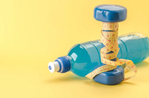 Fitness concept with dumbbell with yellow centimeter and bottle/blue dumbbell with centimeter and bottle on a yellow background. selective focus. Copy space
