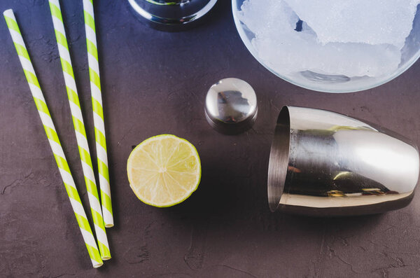 Cocktail shaker, lime, ice and straws / Cocktail shaker, lime, ice and straws on a dark background. Вид сверху
