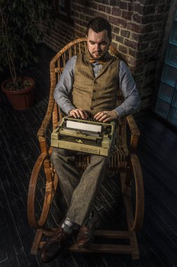 Sallanan sandalye brickwall arka plan üzerinde bir daktilo kullanan genç Hipster adam