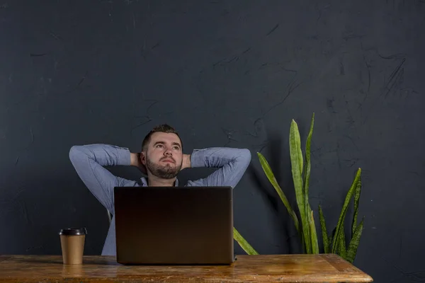 Satisfied with work done. Happy young man working on laptop while sitting at his working place in office. Dark background. Space for Text