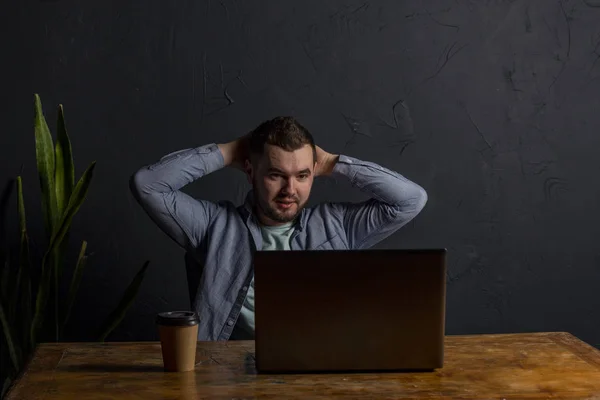 Satisfied with work done. Happy young man working on laptop while sitting at his working place in office. Dark background. Space for Text