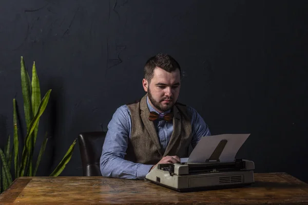 Bearded stylish writer typing on typewriter. Modern writer working on new book in office. Dark background. Space for text