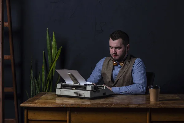 Bearded stylish writer typing on typewriter. Modern writer working on new book in office. Dark background. Space for text