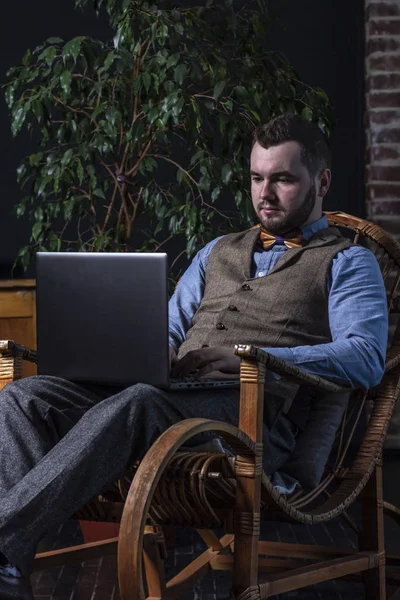Young handsome man with laptop sitting in rocking chair on brickwall background