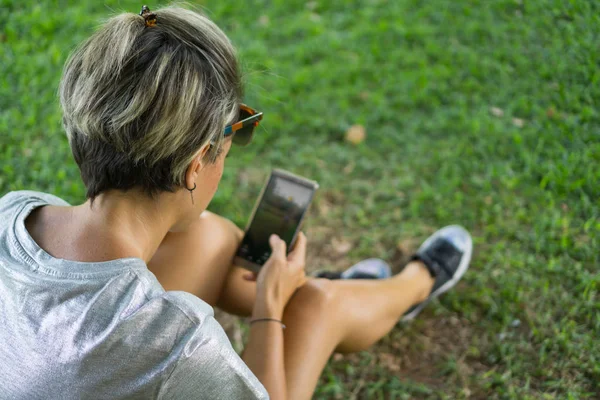 Woman sitting on the lawn with cell phone in a hand. View from back. No logos and brands.