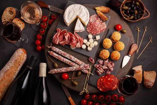 Appetizers table with differents antipasti, cheese, charcuterie, snacks and wine. Mini burgers, sausage, ham, tapas, olives, cheese variety board and baguette over grey concrete background. Top view, flat lay