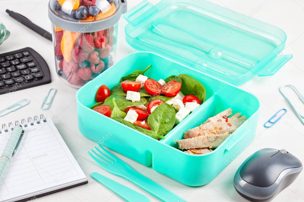 Take away lunch box with fresh salad and tuna fish over the office desk with office supplies. Healthy and balanced lunch at the desk concept