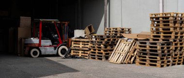 Kırmızı ve gümüş bir forklift bir deponun önünde, yüksek ahşap paletlerin yanına park edilmiş..