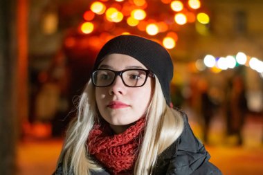 Güzel genç kız bardaklarda gece gece ışıkları fonunda poz. Siyah şapkalı sarışın gülümsüyor.