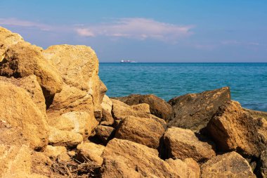 Açık güneşli bir günde Karadeniz manzarası. Kıyıda, taşlar ve kum, ufukta gemiler. Mavi gökyüzü ve güzel bulutlar.