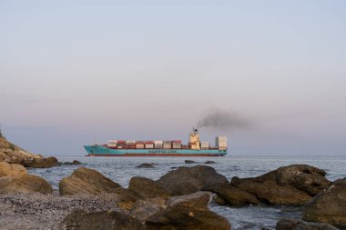Kargo ile ağır bir konteyner gemisi römorkör ile limana girer. Yuzhny Limanı. Ön planda kıyı taşları. Seçici odak. 2019.09.01. Odessa. Ukrayna.