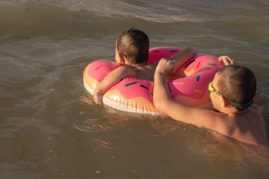 5 yaşındaki bir çocuk 10 yaşındaki abisiyle donut şeklinde şişirilebilir bir çemberle denizde yüzüyor. İki kardeş suya dalar.