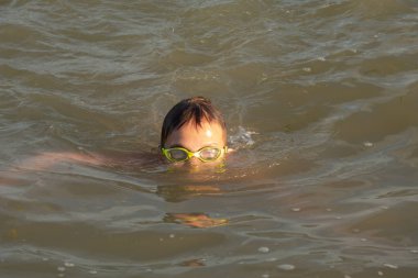 10 yaşında yakışıklı bir çocuk farklı stillerde denizde yüzer. Sırtüstü, kurbağalama, sürünerek.