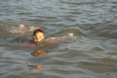 10 yaşında yakışıklı bir çocuk farklı stillerde denizde yüzer. Sırtüstü, kurbağalama, sürünerek.