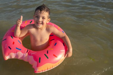 5 yaşındaki bir çocuk donut şeklinde şişirilebilir bir çemberle denizde yüzüyor ve ona benzer bir şey gösteriyor.