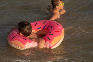 5 yaşındaki bir çocuk 10 yaşındaki abisiyle donut şeklinde şişirilebilir bir çemberle denizde yüzüyor. İki kardeş suya dalar.